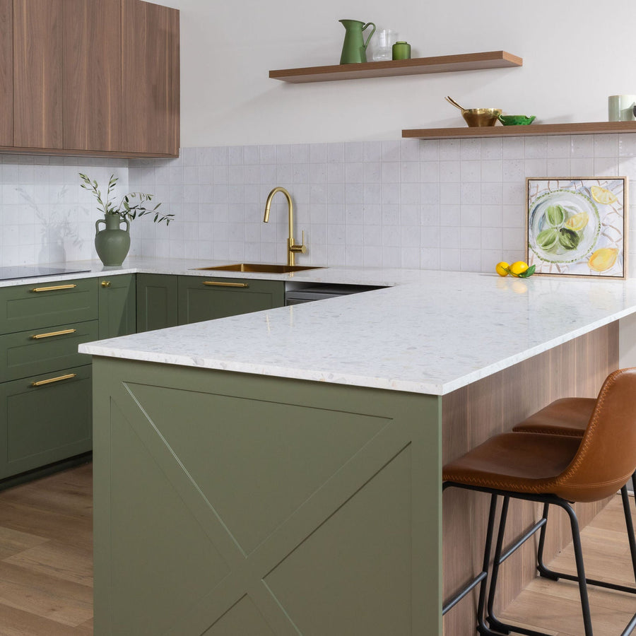 Kitchen with olive green painted cabinet doors, brass handles, a white stone benchtop and timber floating shelves