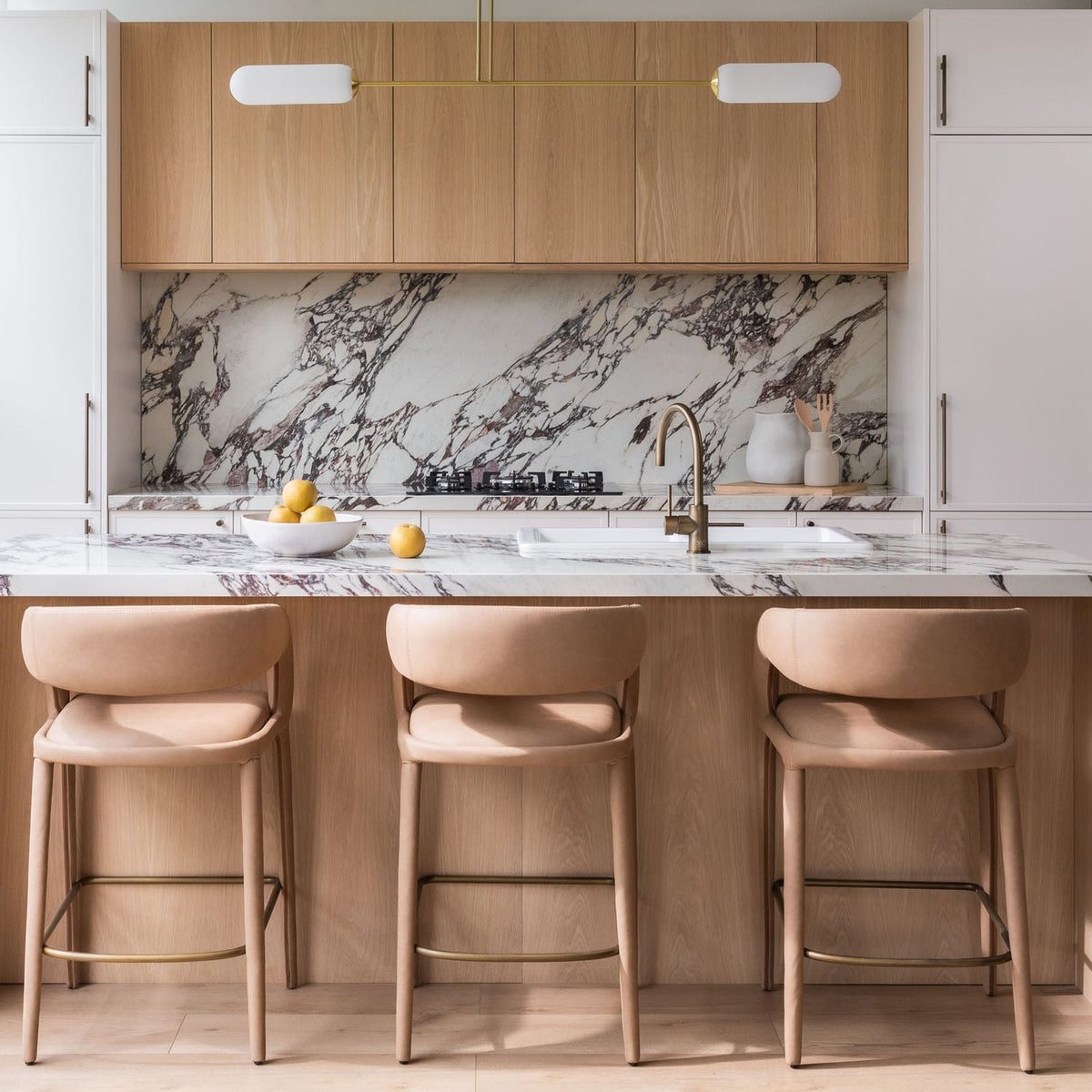Two-tone kitchen in the Customea display with timber-look wall cabinets, white tall doors, a marble splashback and an island with three stools