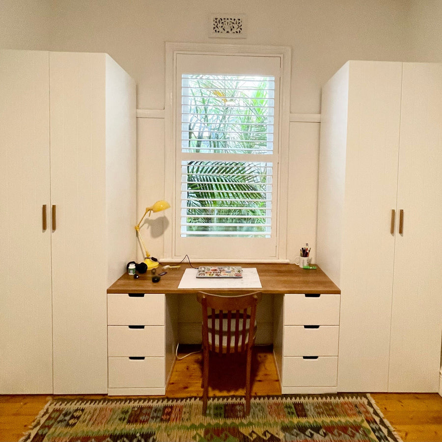 Home office with two IKEA PAX cabinets with white doors either side of a timber desk below a window