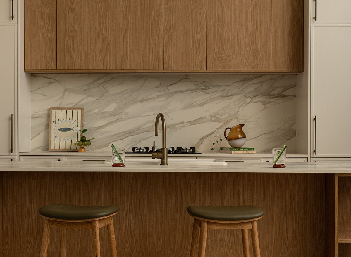 Kitchen with timber cabinets, a marble-look splashback and island benchtop in Caesarstone 506 Mirabel porcelain, two olive leather stools at the island